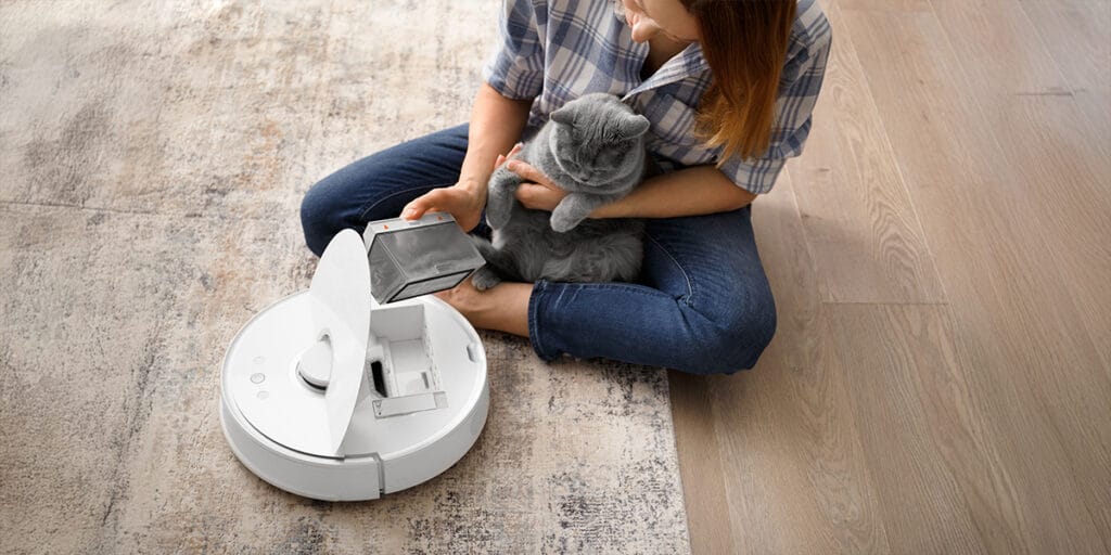 Robot süpürgenin toz haznesi kadının elinde, robot süpürgenin toz haznesi kedi tüyü ile dolu, kedi hazneye bakıyor